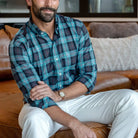 Man wearing Ledbury Blue Albini Olson Plaid shirt seated on leather sofa, paired with white chinos, sleeves rolled