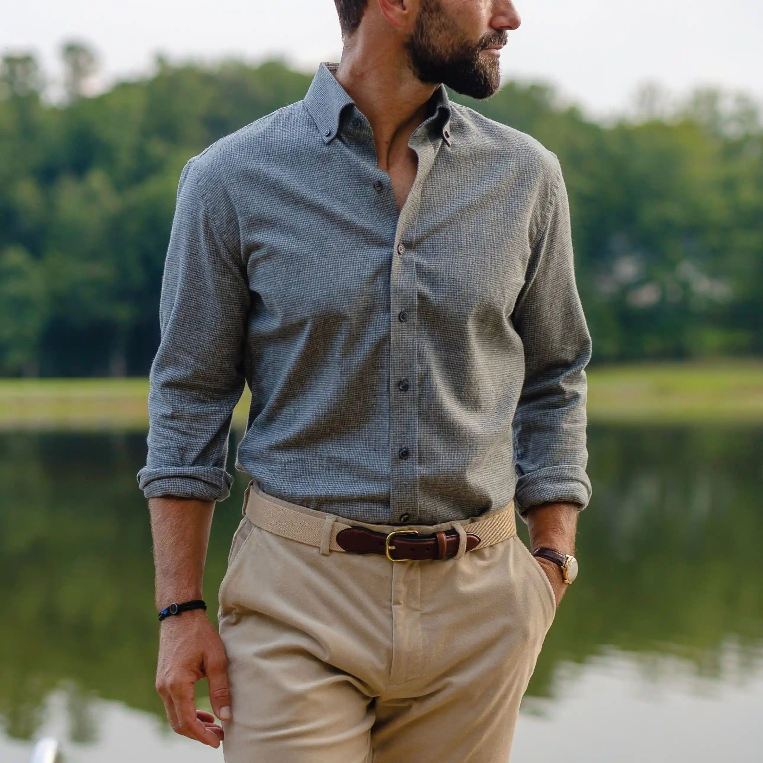 A man wearing a houndstooth grey long-sleeve button-down shirt and beige trousers, standing outdoors near a lake with trees in the background.
