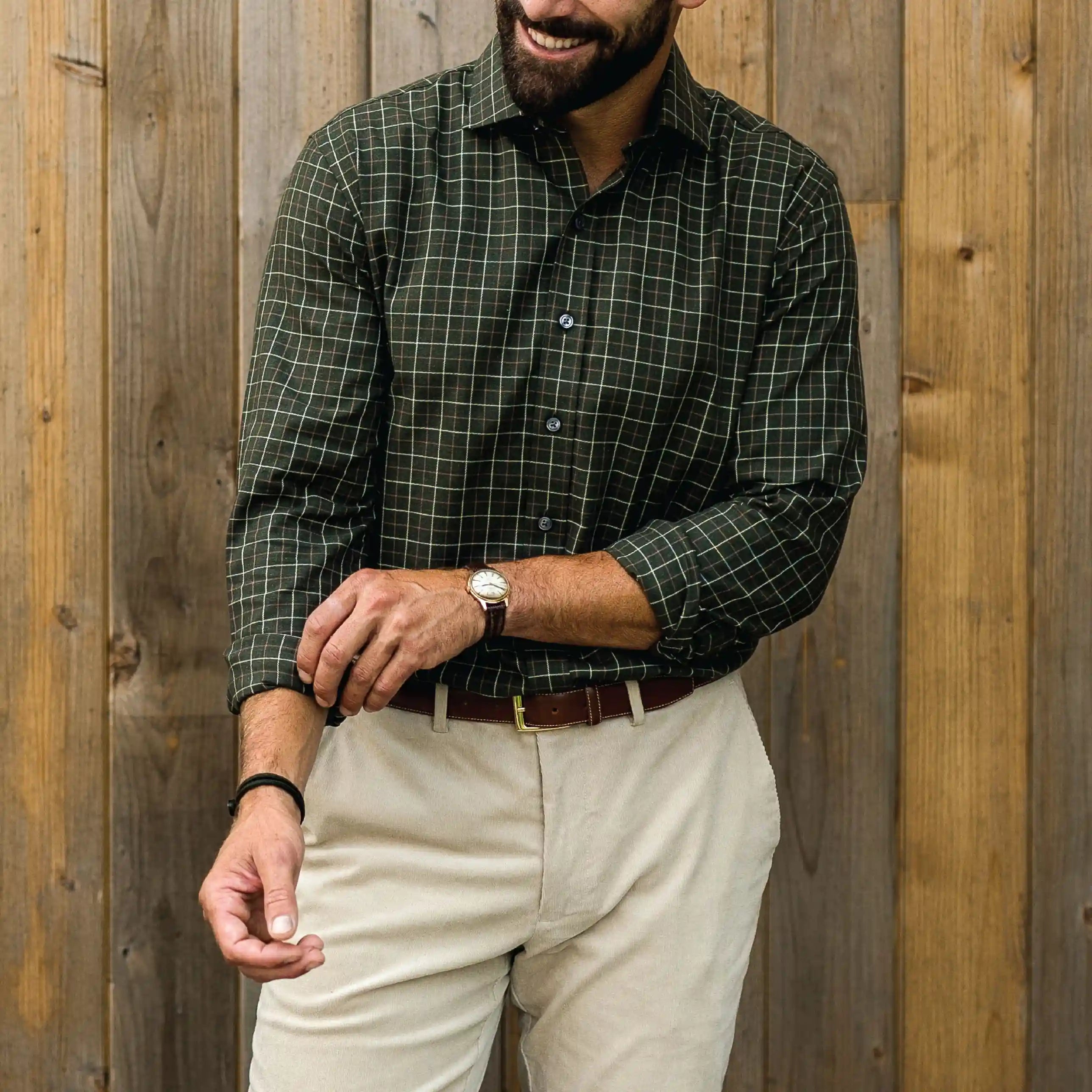 A man wearing a dark green and black plaid long-sleeve button-down shirt with beige pants, standing in front of a vertical wooden plank wall.