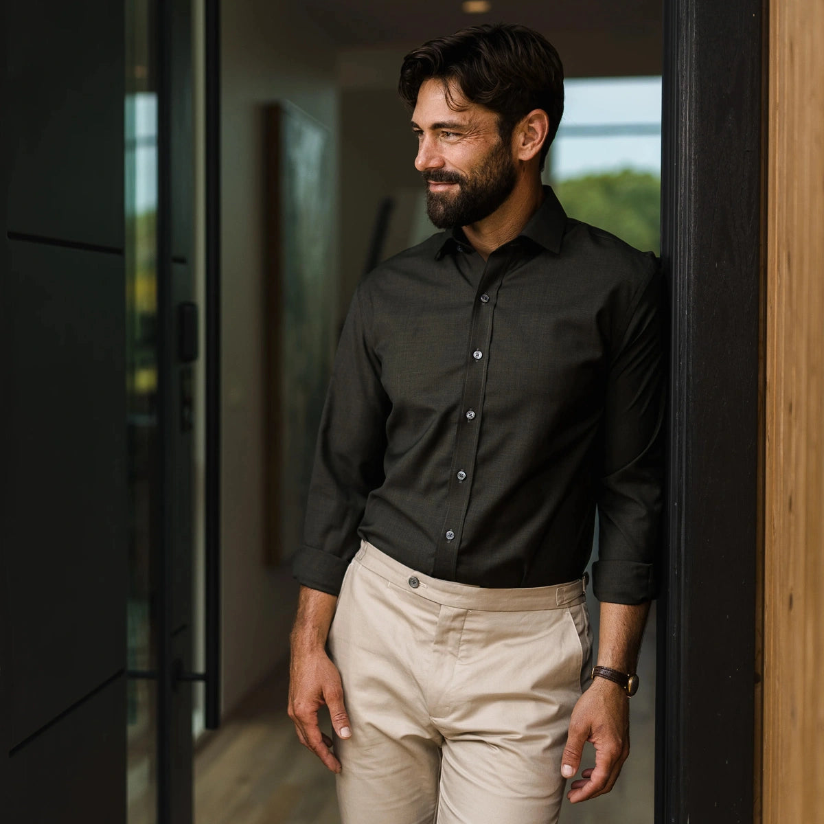 A man wearing a dark green long-sleeve fine Merino button-down shirt and beige trousers, standing in a doorway.