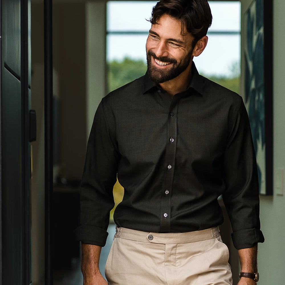 A smiling man wearing a  dark green long-sleeve fine Merino button down shirt and beige trousers, standing in a modern indoor setting with a window in the background.