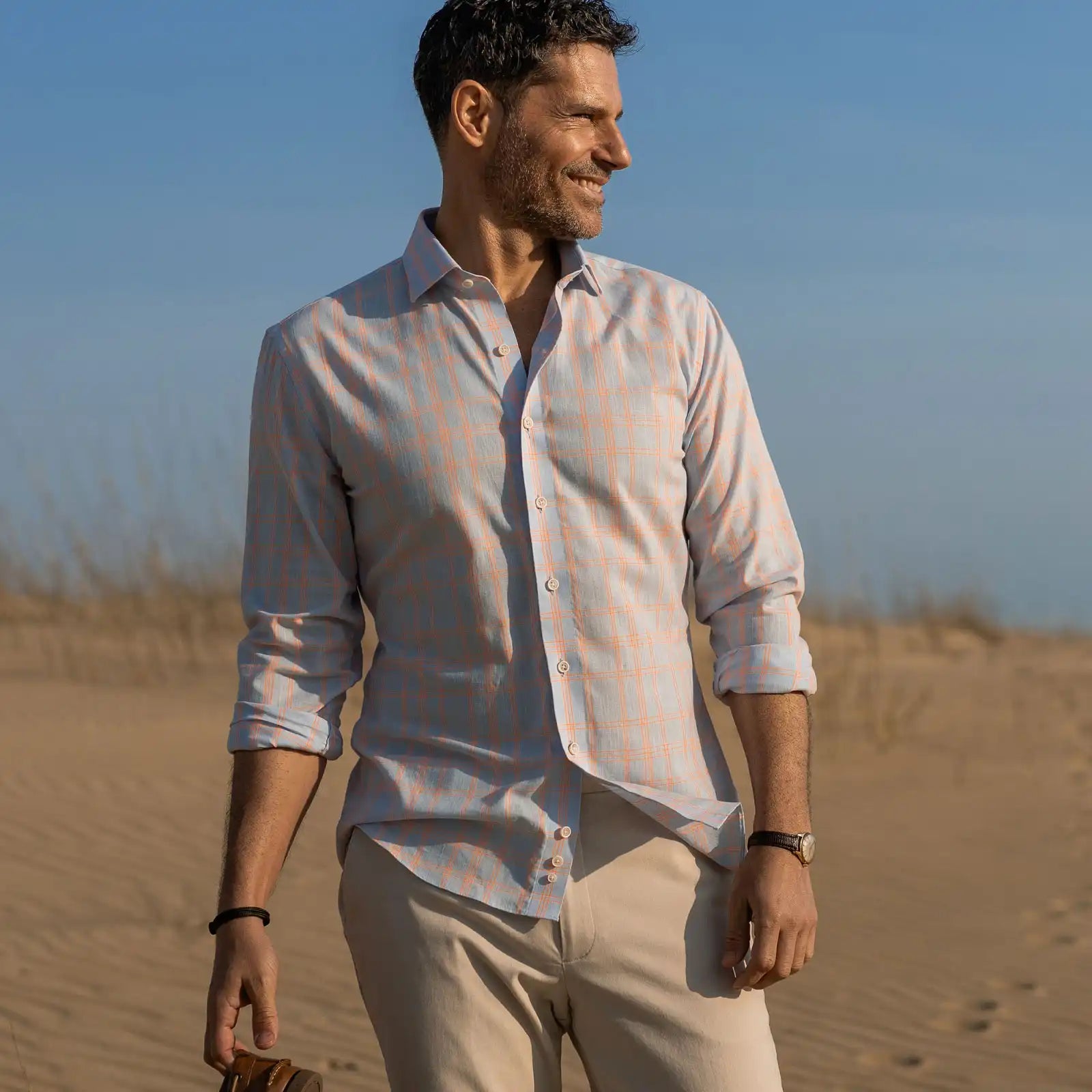 Man standing on a beach wearing a light blue and bright orange checkered shirt and beige pants.