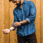 A man wearing a blue denim long-sleeve button-up shirt and dark brown pants, standing in front of a wooden plank wall and adjusting his sleeve.