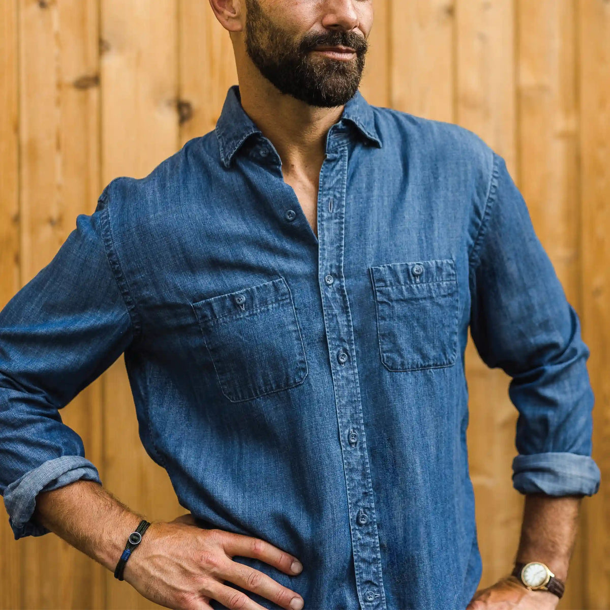 A man wearing a blue denim long-sleeve button-up shirt with two chest pockets, standing in front of a light wood plank wall.