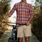 Man wearing a red, blue, and white plaid shirt and beige shorts standing next to a bicycle outdoors.