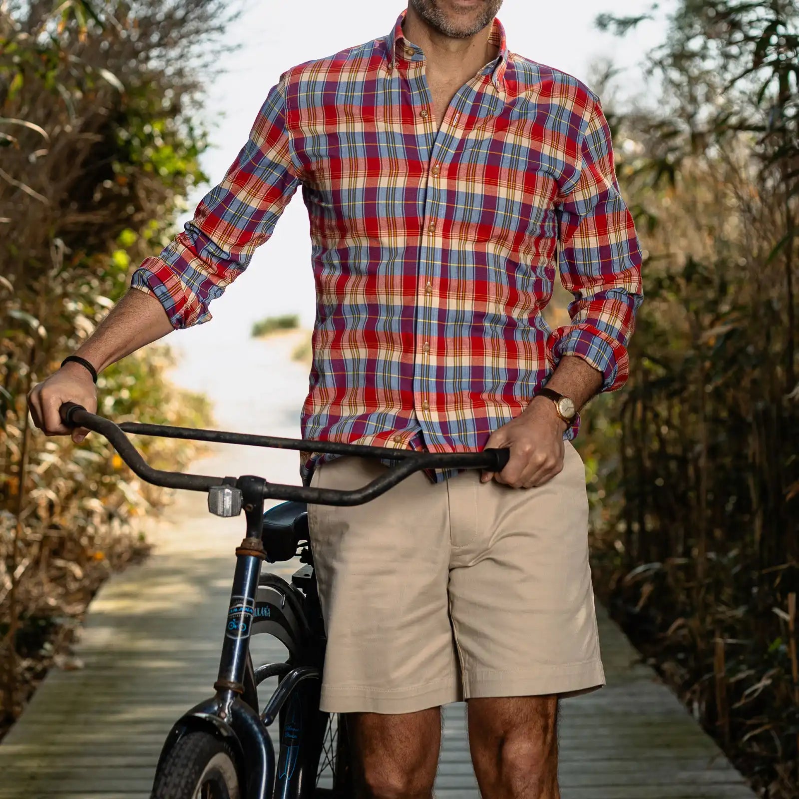 Man wearing a red, blue, and white plaid shirt and beige shorts standing next to a bicycle outdoors.