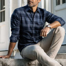 A man wearing a navy blue and gray plaid button-down shirt with beige pants, sitting on concrete steps in front of a white building.