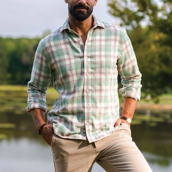 A man wearing a light green and beige plaid long-sleeve button-down shirt, paired with khaki pants, standing outdoors near a body of water with trees in the background.