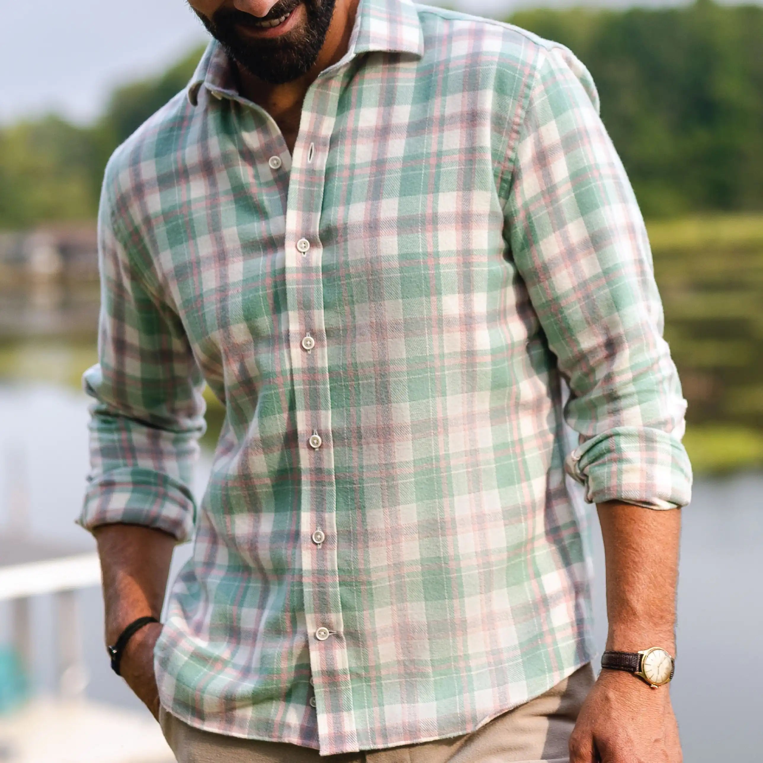 A man wearing a long-sleeve button-up plaid shirt in sage green, pink, and white, with the background showing a blurred outdoor scene of water and trees.