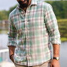 A man wearing a long-sleeve button-up plaid shirt in sage green, pink, and white, with the background showing a blurred outdoor scene of water and trees.