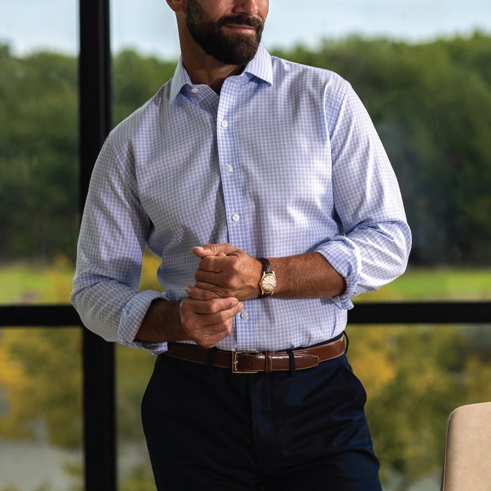 A man wearing a light blue and white checkered long-sleeve button-down shirt, dark navy trousers, and a brown leather belt, standing in front of a window with a blurred outdoor background.