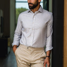 A man wearing a light gray and white micro-check patterned button-down dress shirt with beige trousers, standing in a doorway.