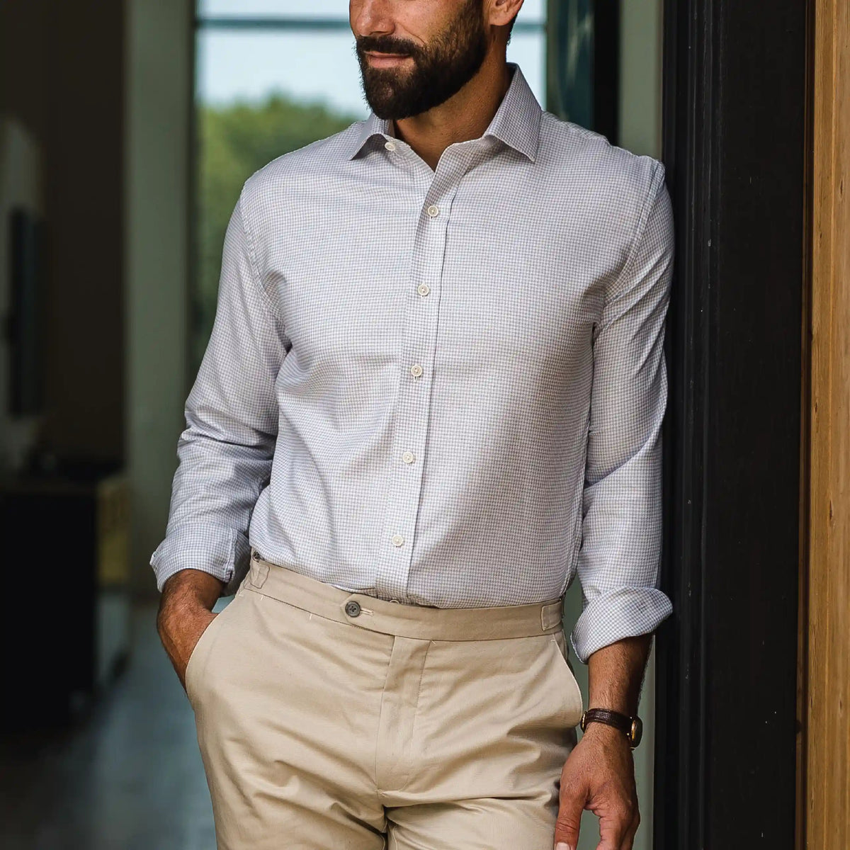 A man wearing a light gray and white micro-check patterned button-down dress shirt with beige trousers, standing in a doorway.