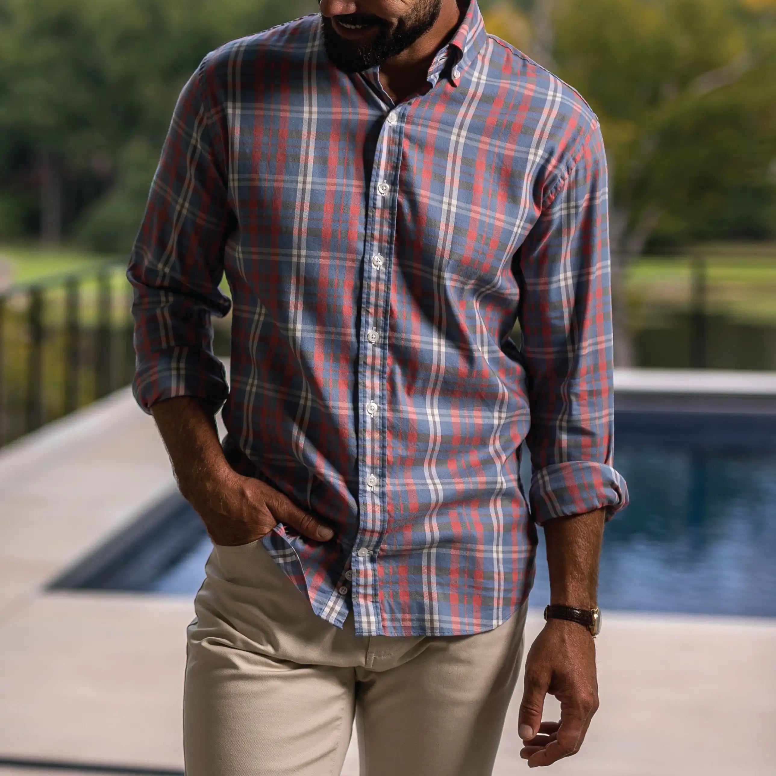 A man wearing a long-sleeve button-down plaid shirt in muted red, blue, and white, paired with khaki pants, standing outdoors near a swimming pool.