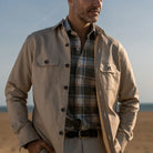 Man wearing a light tan shirt jacket over a plaid shirt with a blurred background
