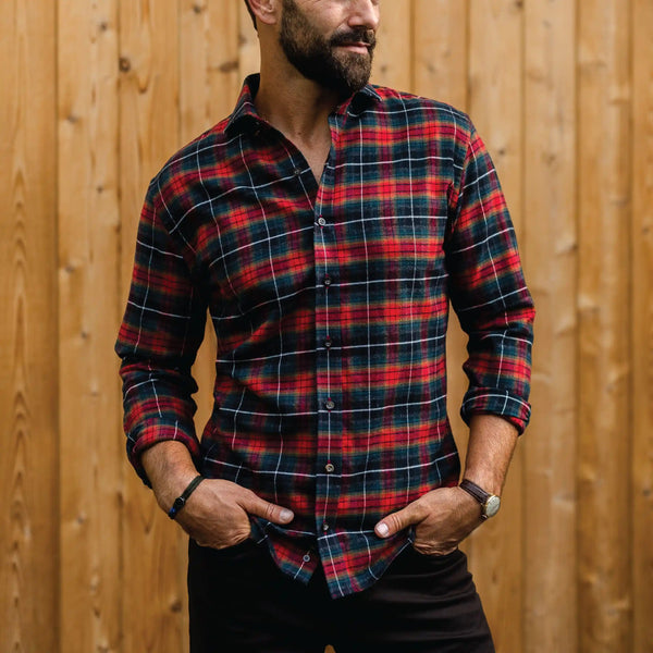 A man wearing a red, black, and white plaid long-sleeve button-down shirt, standing against a vertical wooden plank wall.