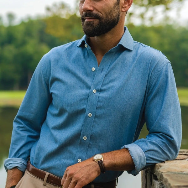 Man wearing a denim look chambray shirt in light blue, standing in front of a lake outdoors. 