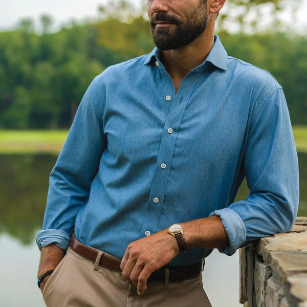 A man wearing a blue long-sleeve button-down shirt and khaki pants, leaning against a stone wall outdoors near water and trees.