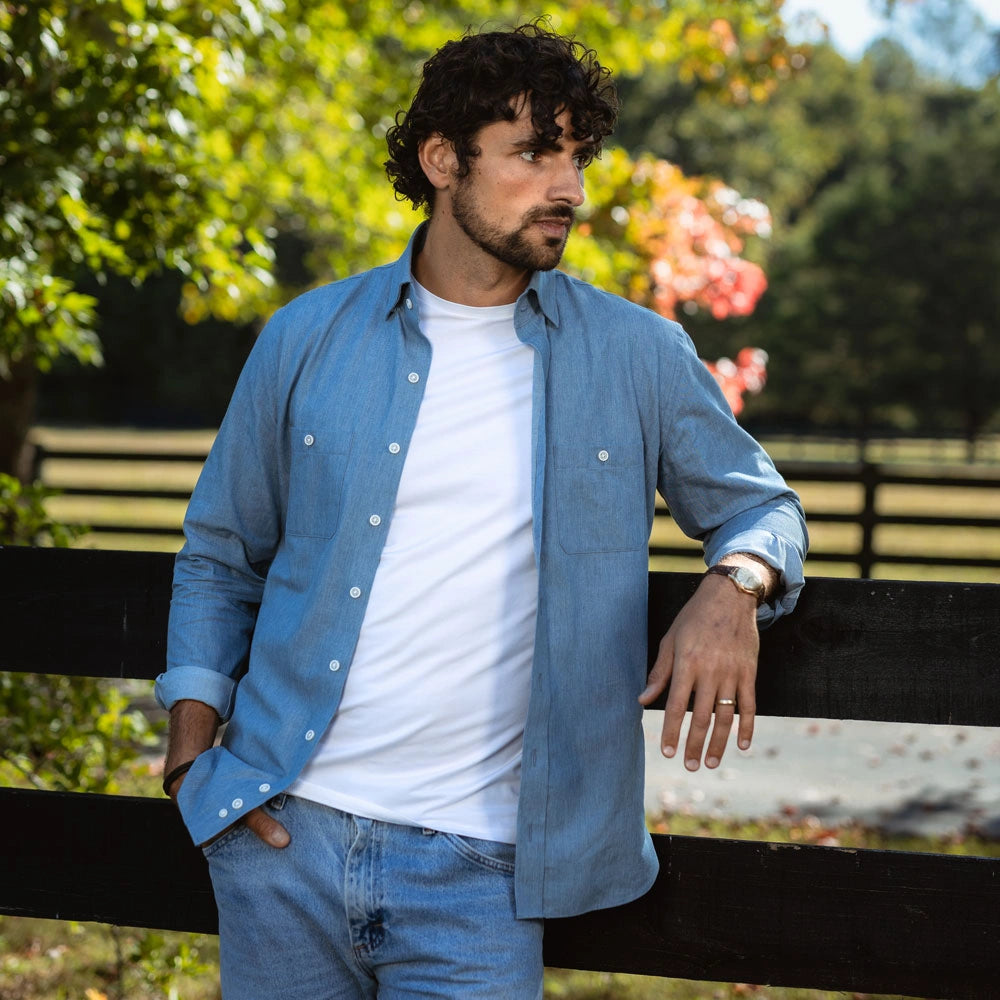 A man wearing a light blue button-down shirt over a white t-shirt and blue jeans, leaning on a black wooden fence in an outdoor park setting with green trees in the background.