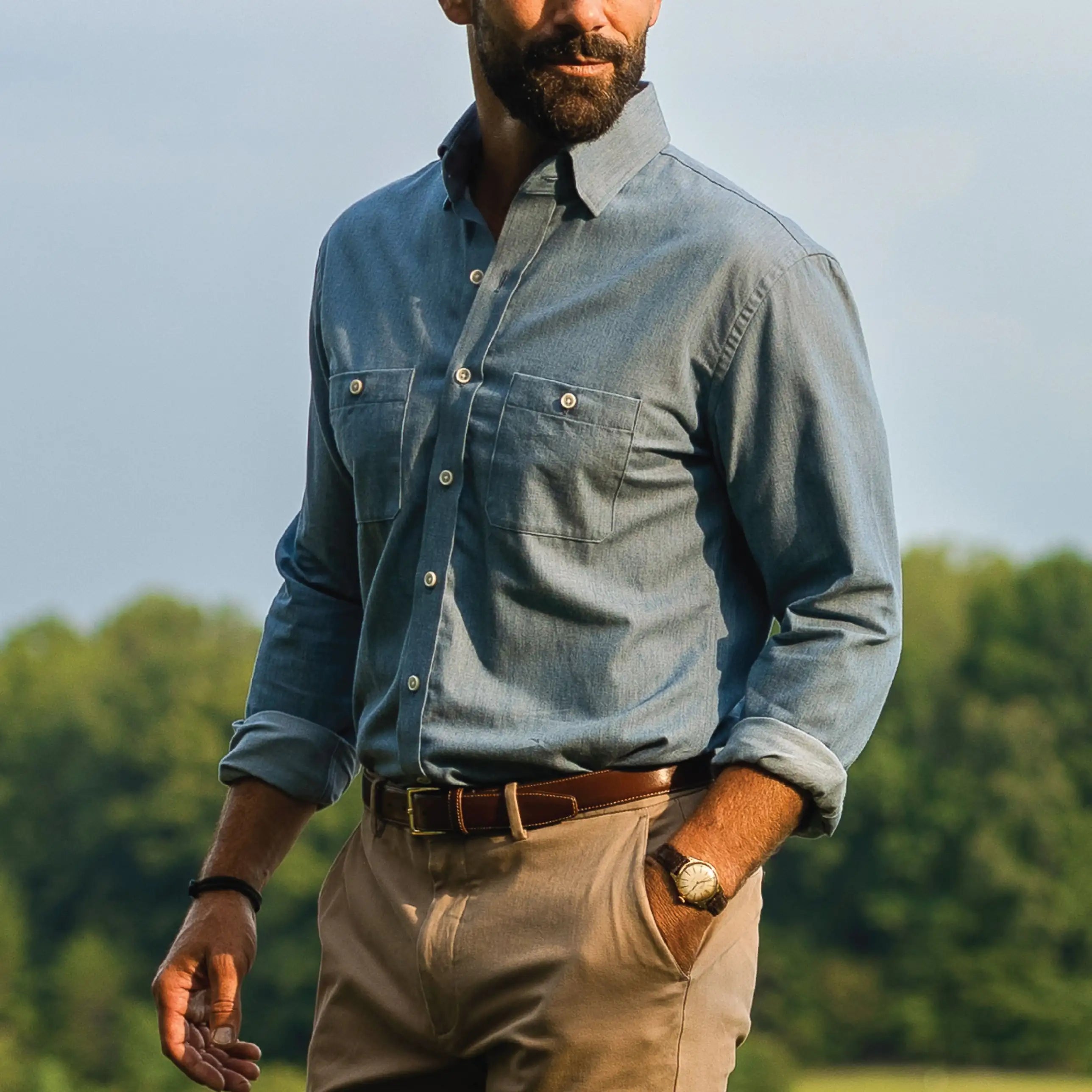 A man wearing a light blue chambray button-down shirt with two chest pockets, paired with tan pants and a brown belt, standing outdoors with trees in the background.