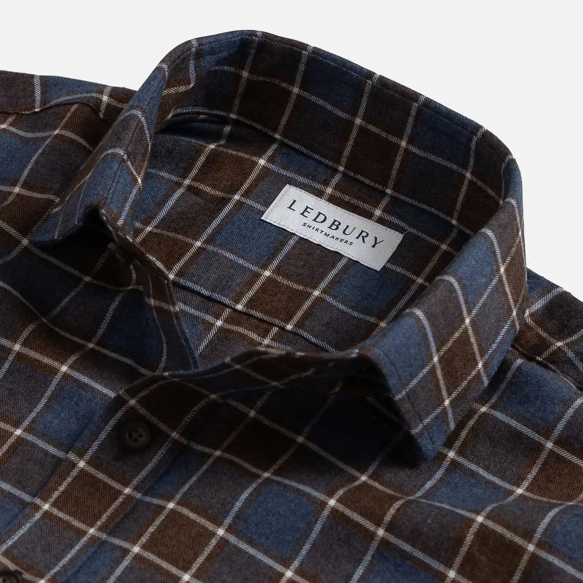 Close-up of a plaid button-down shirt collar with a visible LEDBURY brand label. The fabric is a mix of dark brown, slate blue, and thin white grid lines.