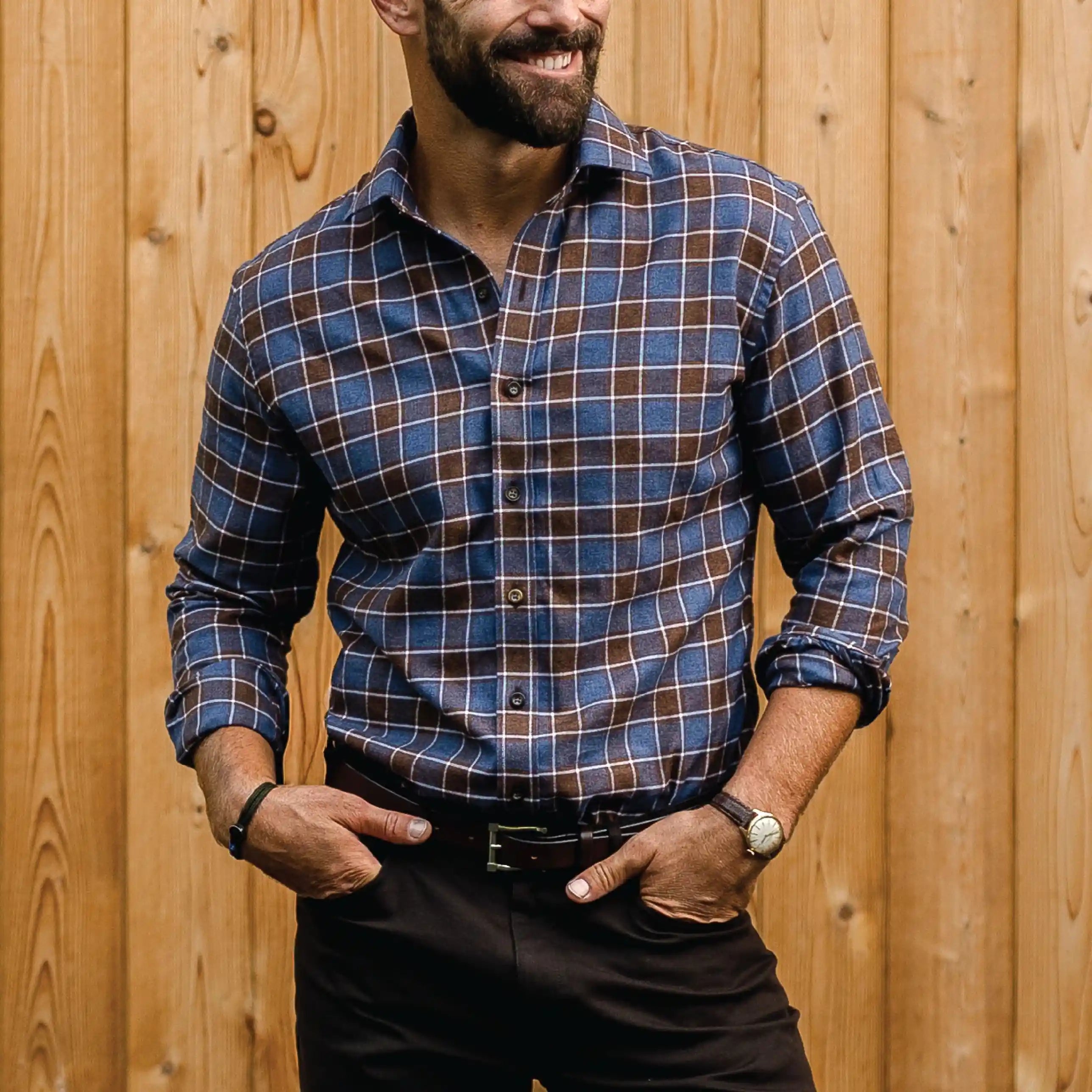 A man wearing a blue and brown plaid long-sleeve button-down shirt, paired with dark brown trousers, standing in front of a vertical wooden plank wall.