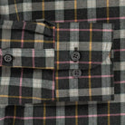 Close-up of a plaid button-up shirt fabric with a dark charcoal base, white and gray checks, and thin yellow and pink stripes. The shirt features a chest pocket with two dark buttons and a matching buttoned cuff.