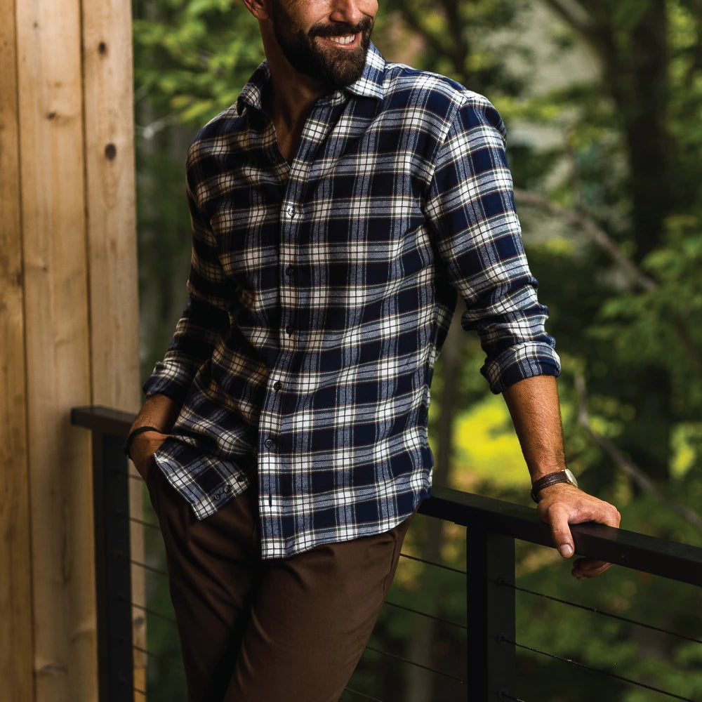 A man wearing a navy blue, white, and gray plaid button-down shirt and brown pants, leaning on a black metal railing on a wooden deck with a forest background.