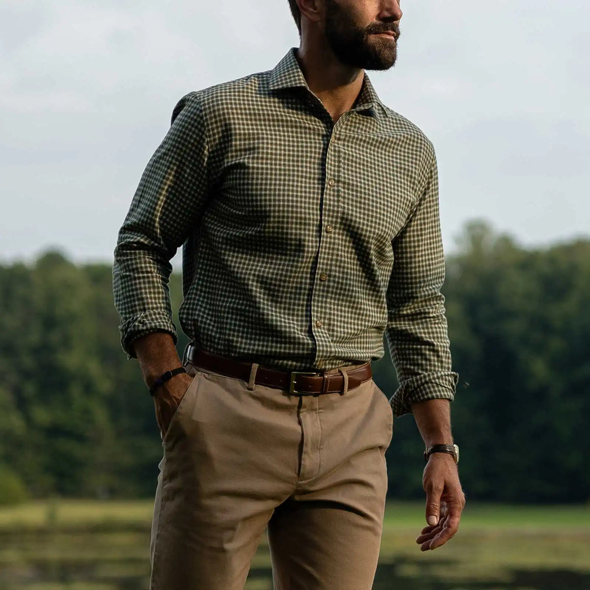 A man wearing a green checkered long-sleeve button-down shirt, khaki pants, and a brown belt, standing outdoors in a grassy field with trees in the background.