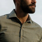 Close-up of a man wearing a green and beige gingham check button-down shirt, with a beard and looking off-camera against a pale sky.