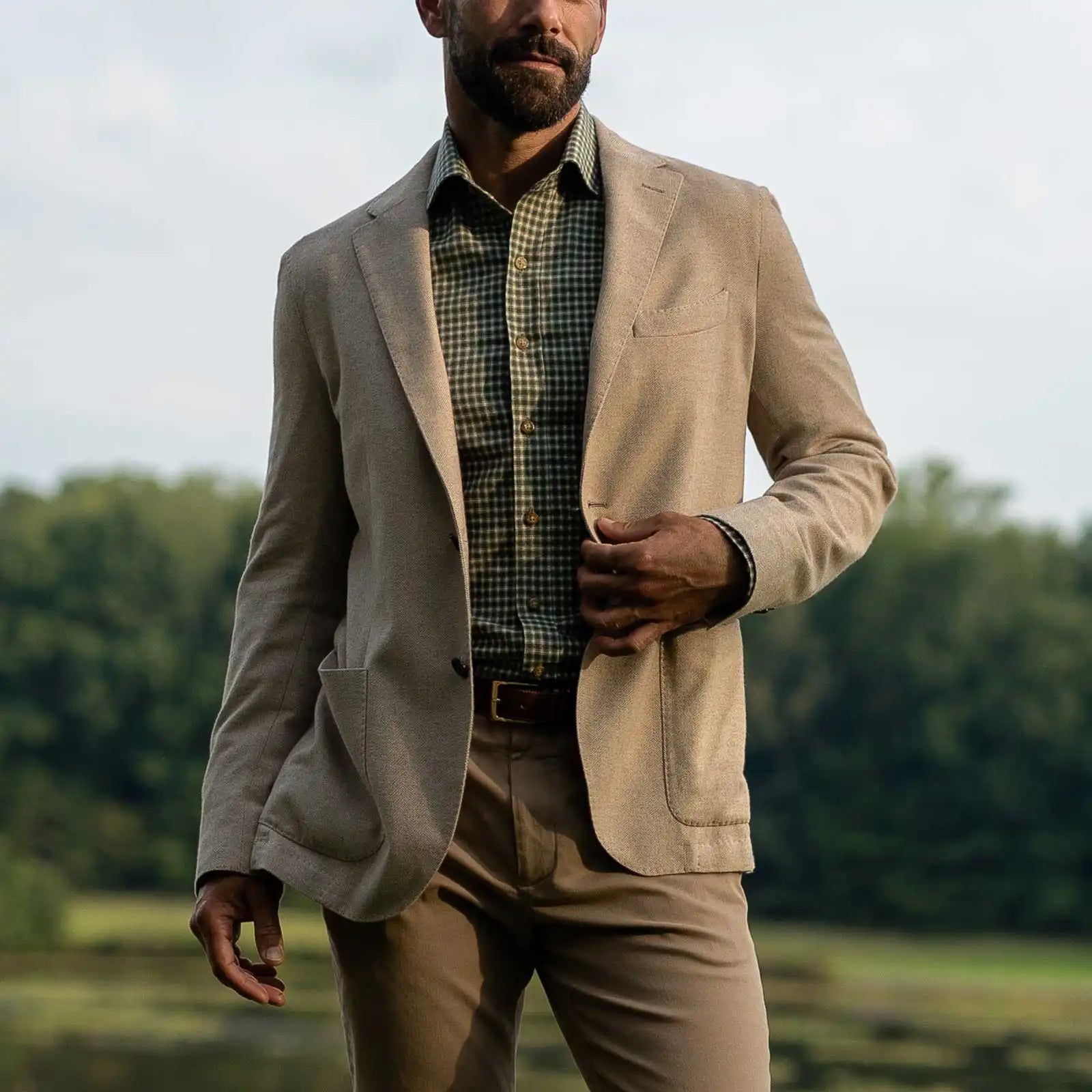 A man wearing a light beige blazer over a green checkered shirt, paired with matching beige trousers, standing outdoors in front of a blurred green landscape.