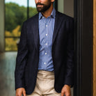 A man wearing a dark navy textured blazer over a blue and white striped button-down shirt, paired with beige trousers, standing in a doorway.