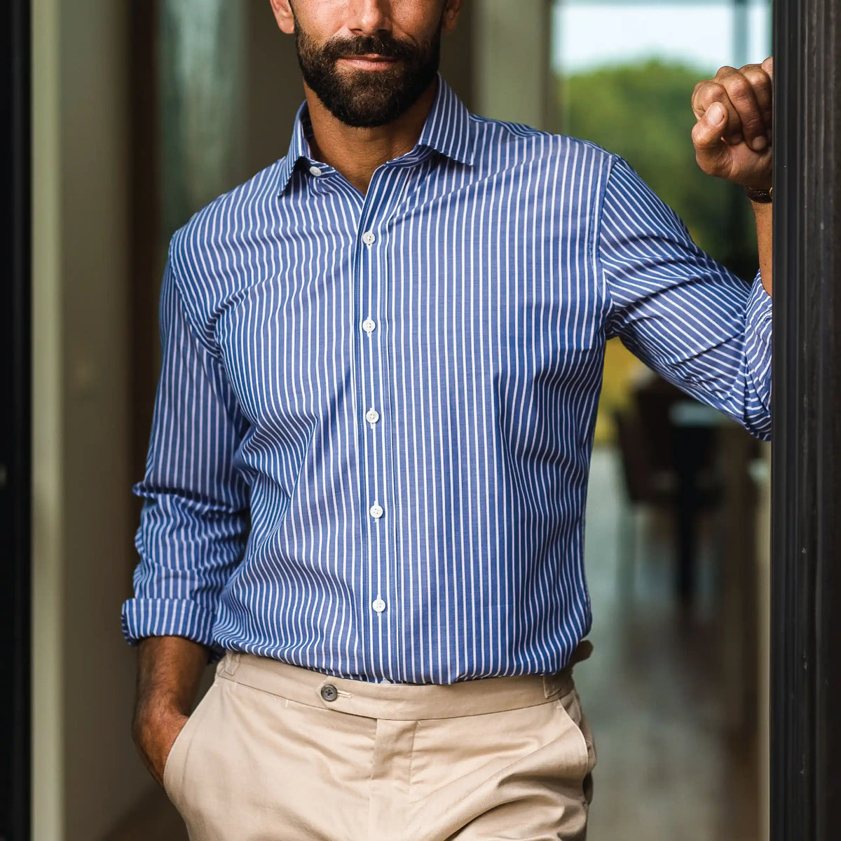 A man wearing a blue and white vertically striped button-down dress shirt with beige trousers, standing in a doorway.