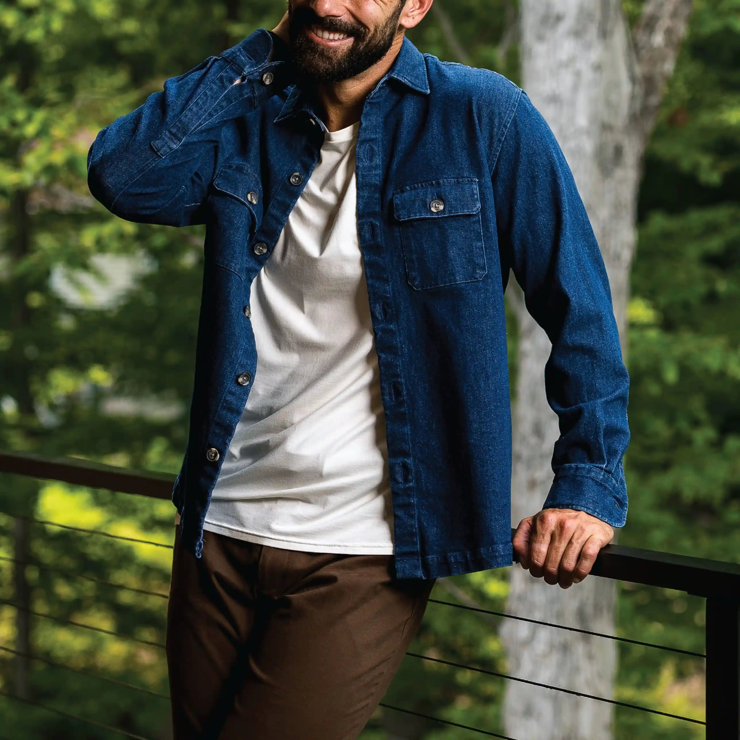 A man wearing a blue denim button-up shirt over a white t-shirt and brown pants, leaning on a railing with a forest background.