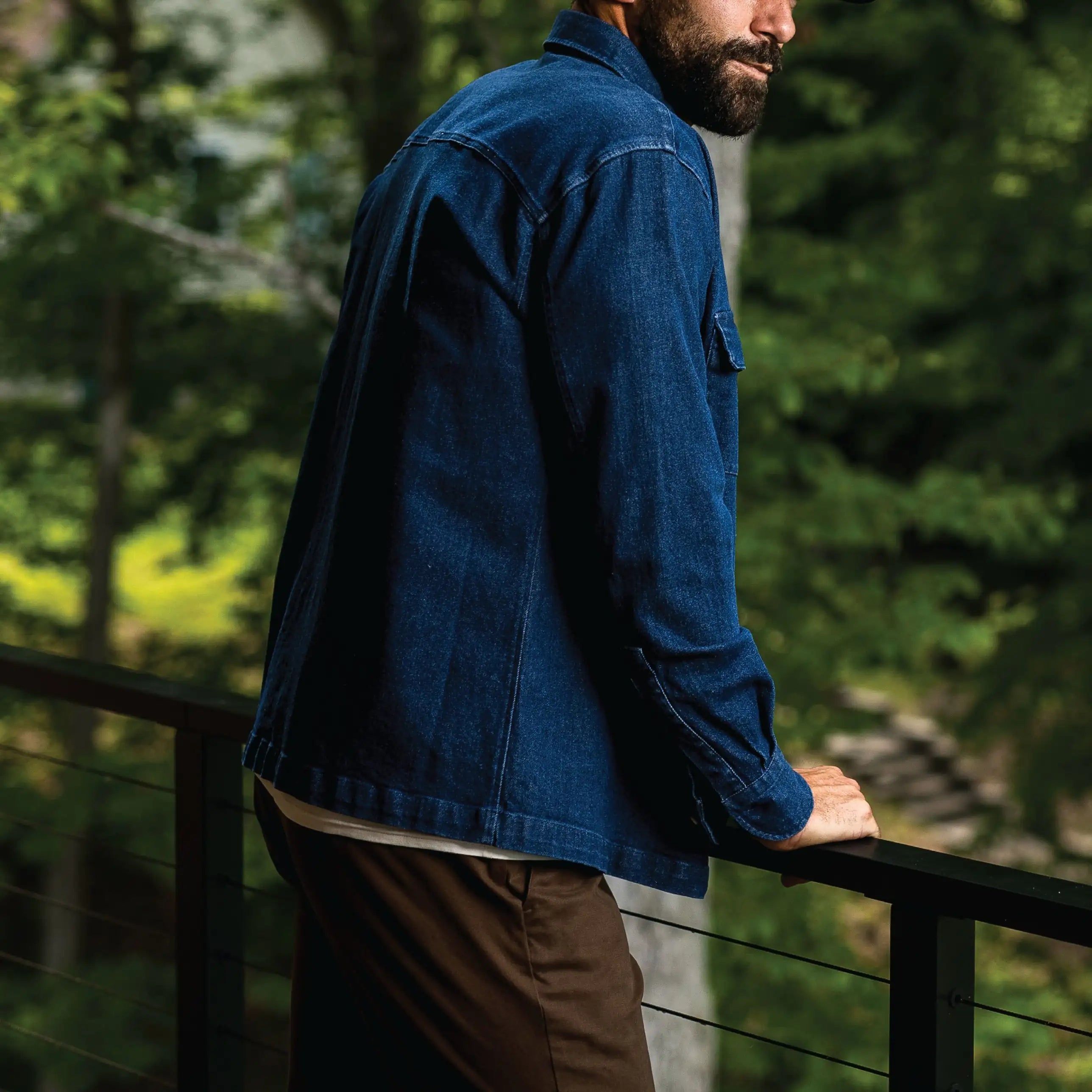 A man wearing a dark indigo blue denim jacket and brown pants, standing on a balcony with a forest background.