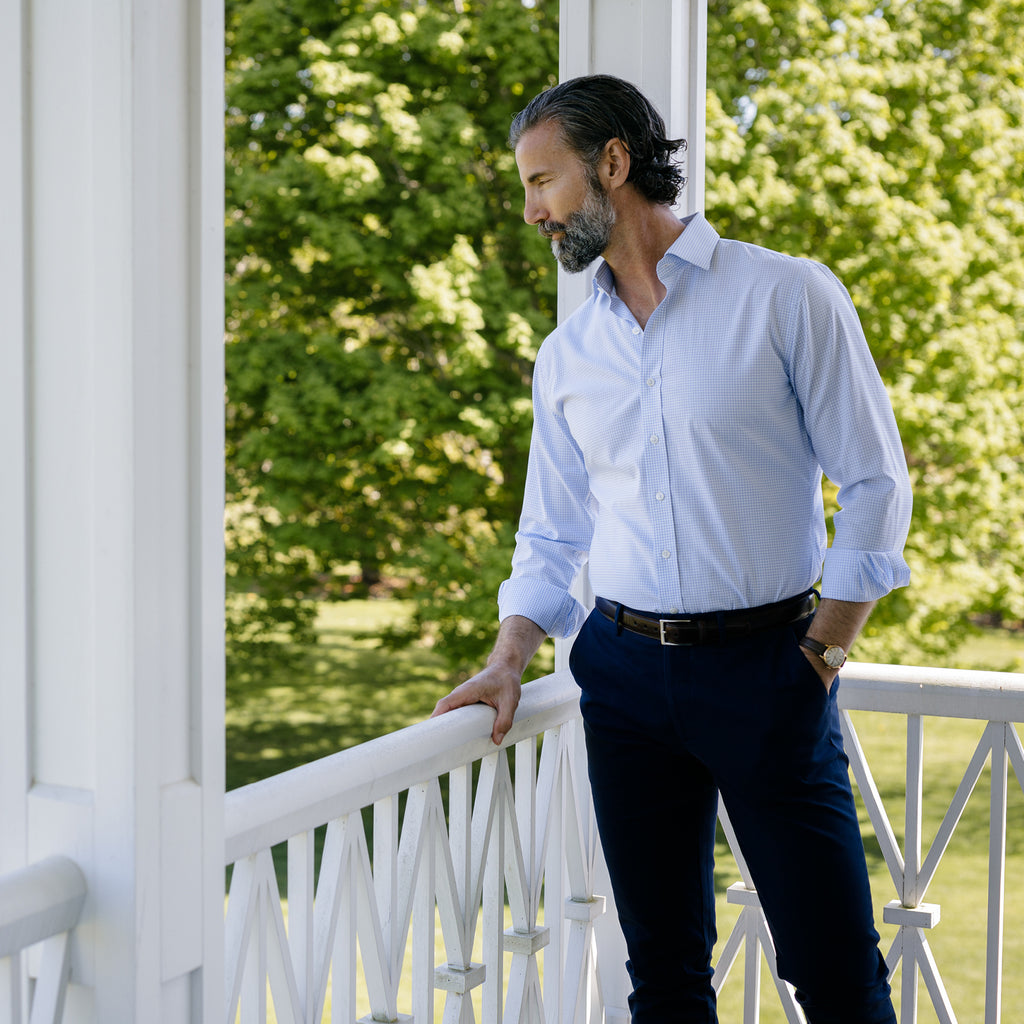 Male model standing on a deck looking over a garden. He is wearing a light blue dress shirt and some navy chinos. 