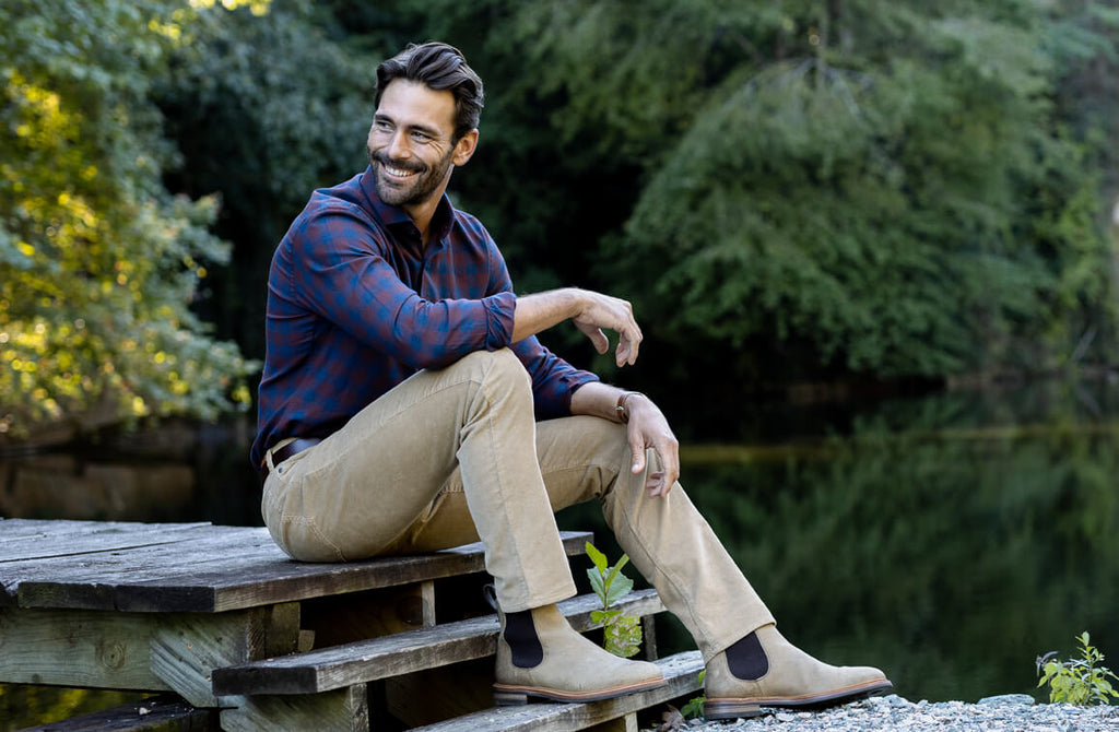 Male model sitting on a lake dock wearing a button down navy and dark red plaid shirt