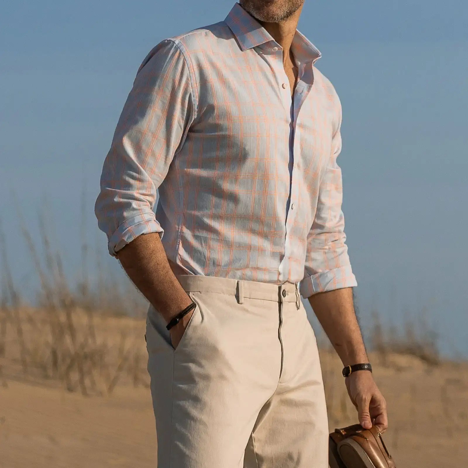 Man standing on a beach wearing a light blue and bright orange checkered shirt and tan chinos