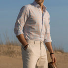 Man standing on a beach wearing a light blue and bright orange checkered shirt and tan chinos