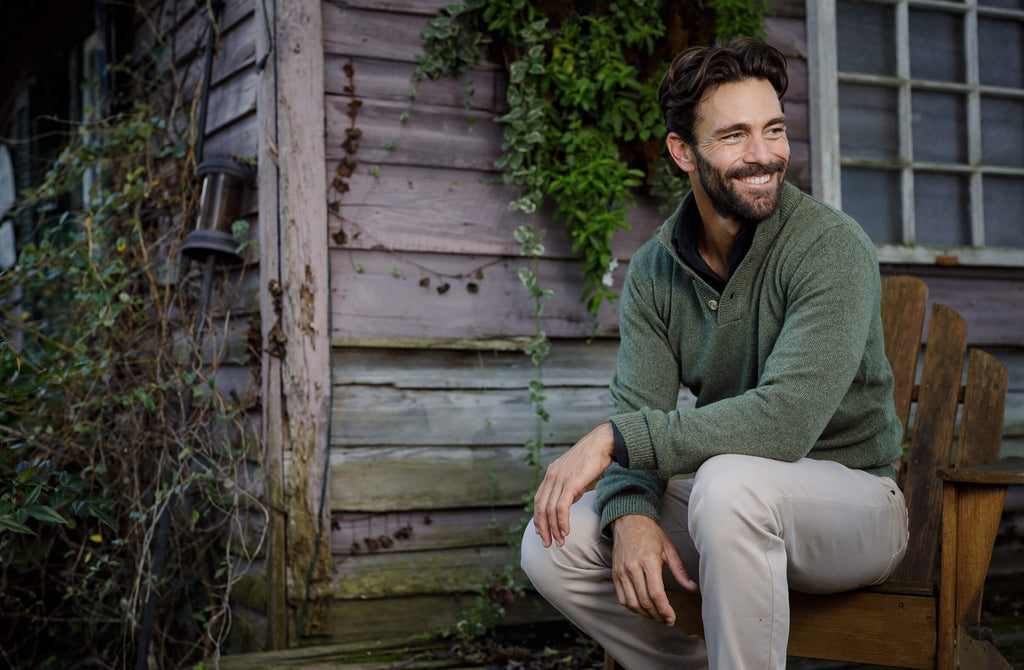 Male model wearing a green crewneck sweater. He is sitting in an adirondack chair outside a rustic house. 