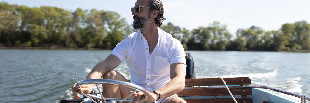Image of a male model driving a boat on a lake. He is wearing a short sleeve white oxford shirt.