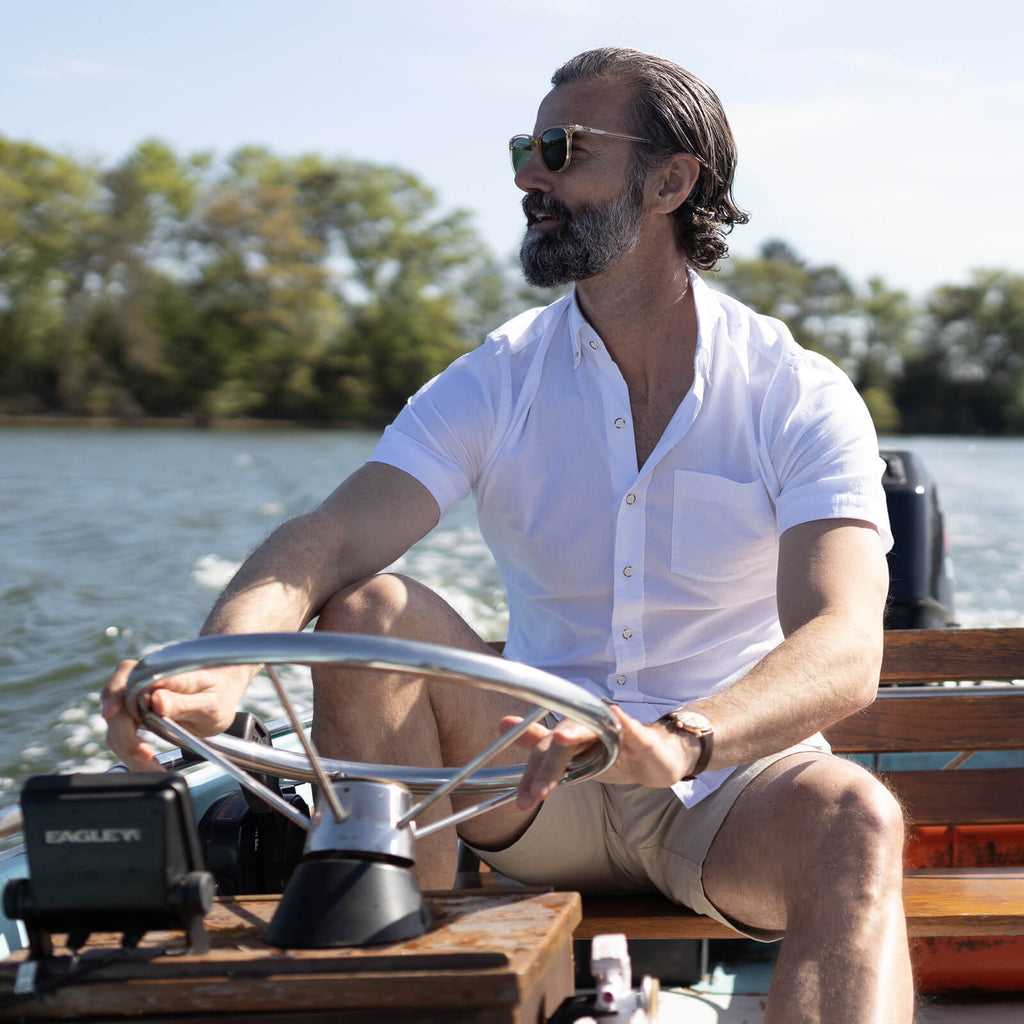 Image of a male model driving a boat on a lake. He is wearing a short sleeve white oxford shirt.