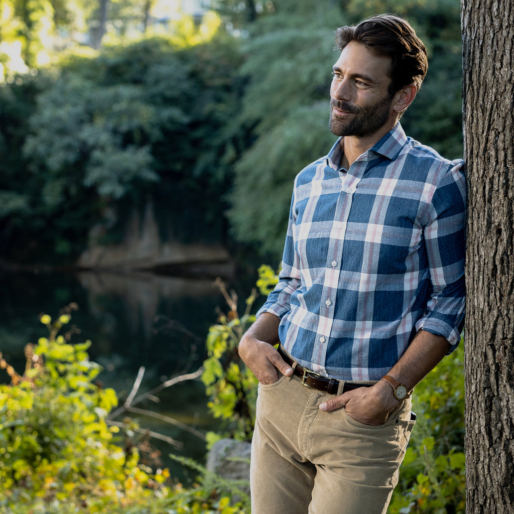 Image of a male model leaning up against a tree wearing a men's blue, pink and white summer weight plaid shirt.