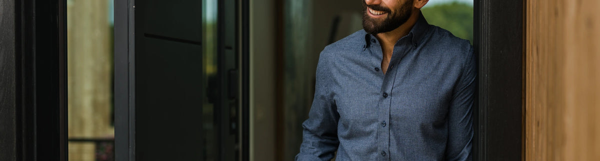 A man wearing a blue button-down shirt stands in a doorway, smiling. The background shows a modern interior with dark glass panels and warm wood trim.