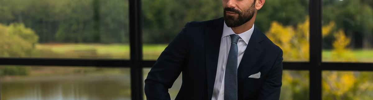A man with a beard wearing a dark navy suit, white dress shirt, and blue tie, standing in front of a large window with a blurred outdoor background of green trees and yellow foliage.