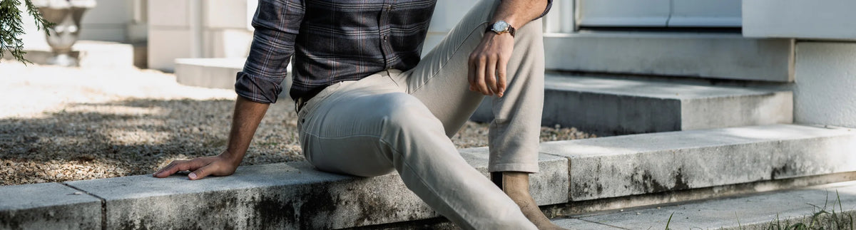 A man sitting on outdoor concrete steps wearing light beige trousers, a dark plaid shirt, and a wristwatch.