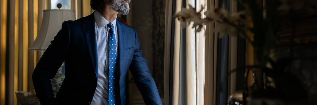 Image of a male model standing in front of a window. He is wearing a blue dress shirt, tie, and blue sport coat.
