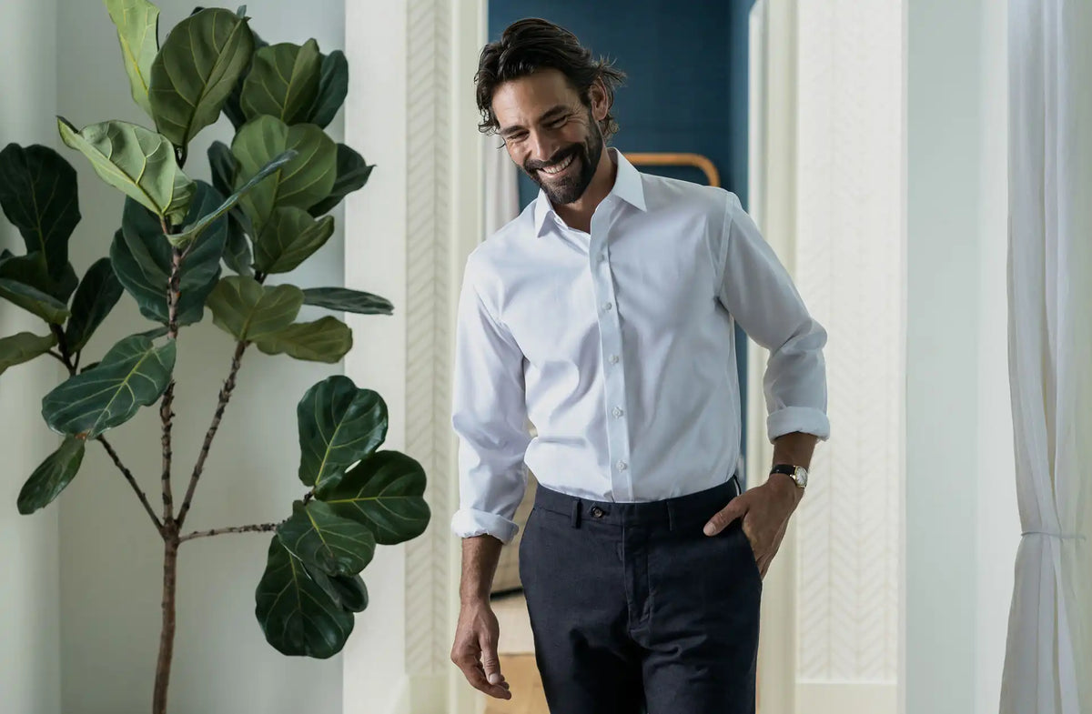 Man wearing a white dress shirt and dark pants standing indoors with a plant to his left.