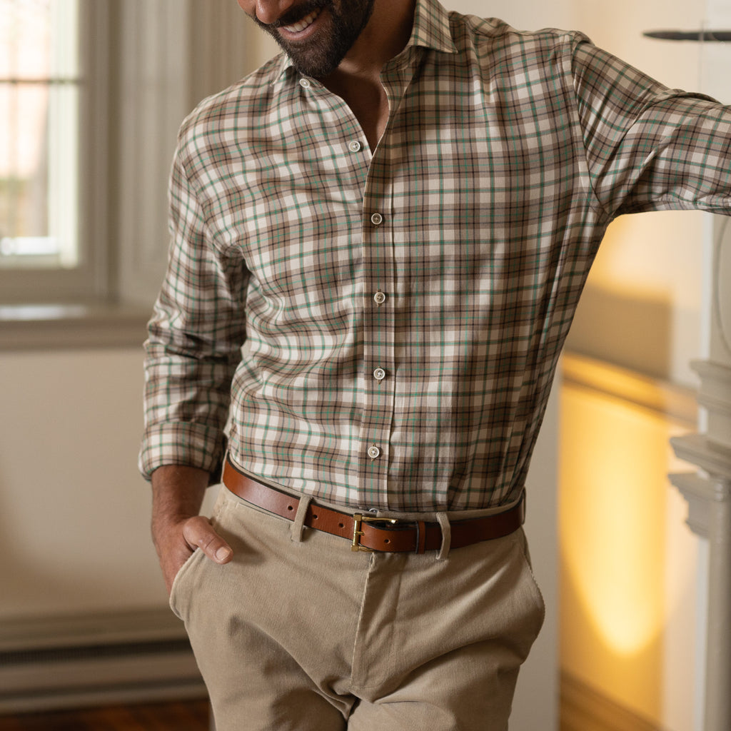 A man smiling indoors leaning against a fireplace with focus on his brown, ivory, beige, and green Bradford plaid shirt.