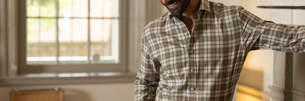 A man smiling indoors leaning against a fireplace with focus on his brown, ivory, beige, and green Bradford plaid shirt.